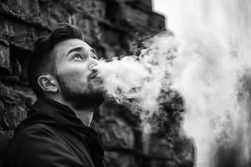 Vape man. Photo of a handsome young white guy with modern haircut in the black clothes vaping and letting off steam from an electronic cigarette near old destroyed brick wall. Black and white photo.