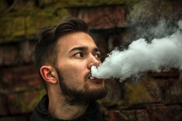 Vape man. Photo of a handsome young white guy with modern haircut in the black clothes vaping and letting off steam from an electronic cigarette near old destroyed red brick wall. Lifestyle.