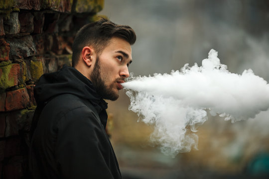 Vape man. Photo of a handsome young white guy with modern haircut in the black clothes vaping and letting off steam from an electronic cigarette near old destroyed red brick wall. Lifestyle.