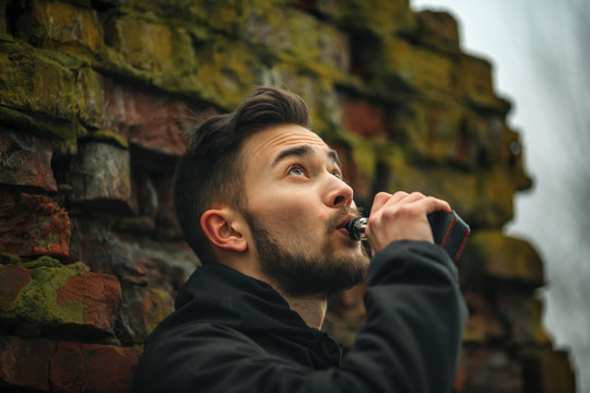Vape man. Photo of a handsome young white guy with modern haircut in the black clothes vaping and letting off steam from an electronic cigarette near old destroyed red brick wall. Lifestyle.