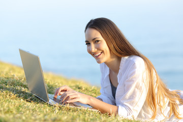 Happy woman using a laptop on the grass looking at you