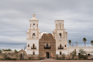Fototapeta na wymiar Tucson, Arizona, USA - January 9, 2018: White and brown front facade of Historic San Xavier Del Bac Mission under heavy gray, white cloud deck. Some greenish desert plants in front.