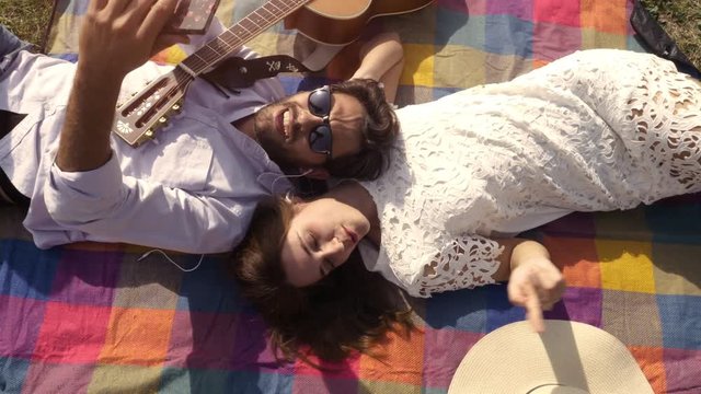 Happy lovely young couple lying on colorful blanket in the park taking selfies smartphone romantic with guitar sunglasses beautiful attractive girl top view rotating camera slow motion