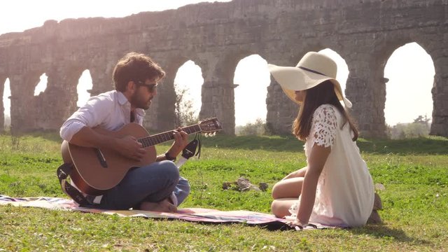 Happy young lovely couple sitting lying on a blanket in the grass in front of ancient roman aqueduct ruins in parco degli acquedotti park in rome romantic play guitar sing beautiful girl with hat