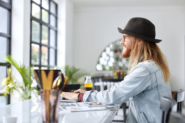 Smiling male freelancer has specific appearance, keyboards on laptop computer, connected to wireless internet, sit at table with lunch against modern kitchen or cafeteria interior. Technology, people