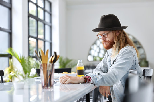 Concentrated bearded male in trendy hat reads magazine while sits at kitchen table with dinner, has serious expression. Hipster guy studies popular literature for creating new project. People, style