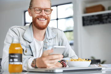 Image of cheerful ginger male with thick beard and mustache wears denim jacket, happy to recieve message from best friend on cell phone, has dinner break after work in canteen, smiles happily