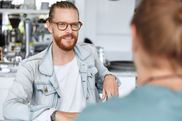 Obraz premium Stylish trendy looking young male freelancer in eyewear and denim jacket meets with his partner for discussing some details and work conditions, have informal meeting together at cozy coffee bar