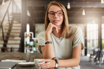Attractive female freelancer wears trendy spectacles and casual t shirt, being deep in thoughts as drinks aromatic coffee, earns money in internet, thinks about something. Creative woman in cafe