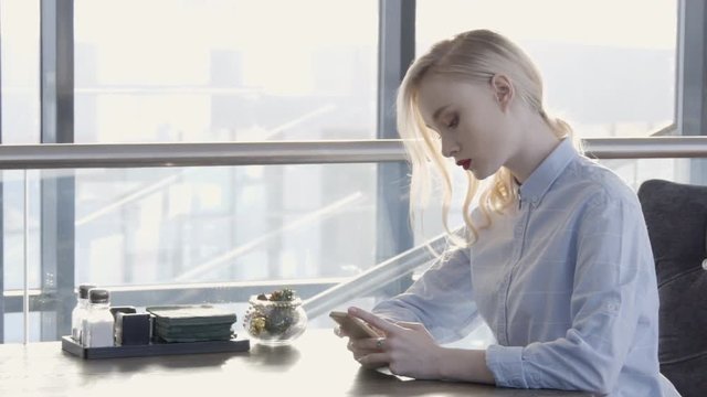 Lonely young blond woman is sitting in a cafe with a phone. Beautiful woman at table and waiting for an order and at same time writing a text message on a smartphone over Internet.