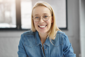 Close up portrait of positive blonde woman in round spectacles, denim jacket, being satisfied with work results, sits in spacious cabinet, expresses positive emotions. People, facial expressions