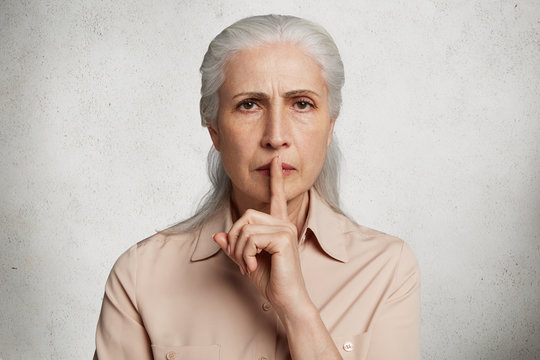 People, body language concept. Old female model keeps fore finger on lips, asks to be silent as has headache, dressed in elegant shirt, isolated over white background. Pensioner gestures indoor