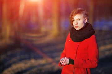 Portrait of young cute girl with a stylish haircut in a red coat and black scarf at the neck, posing in bright sun on a blurred background of trees in Park