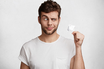 Amazed young brunet male holds condom, raises eyebrow and looks at safety product in bewilderment,...