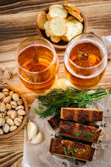 two glasses of light beer, pistachios and croutons on a wooden table.