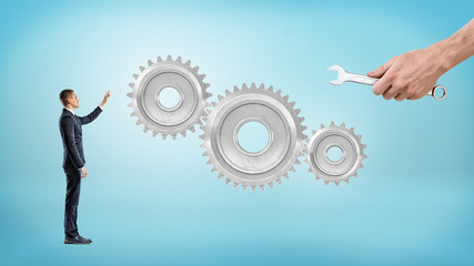 A small businessman points to giant interlocked gears while a big male hand holds a wrench near them.