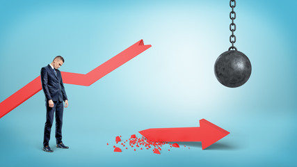 A sad businessman stands near a broken red statistic arrow with a wrecking ball hanging nearby.
