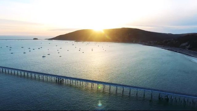 Aerial Flyover - Ocean sunset with sailboats, a pier and mountains
