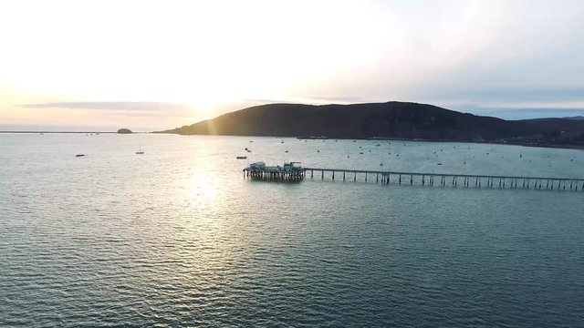 Aerial Drone Flyover - Pacific ocean bay at sunset. In the scene is a pier, sailboats, mountains and sky.