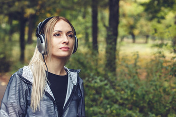 Portrait of young attractive blonde woman on a city park, listening to music on headphones