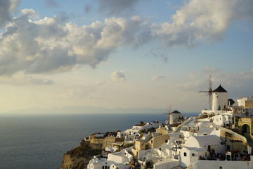 Fototapeta premium Beautiful evening light scene of Oia white building townscape and windmill along island mountain, vast Aegean sea, abstract cloud and blue sky background