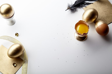 Fototapeta premium Easter holiday composition with eggs painted golden, yolk in egg stand, black feather and golden ribbon. White background, copy space