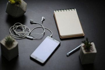 Black mobile phone with white earphones, small succulents in concrete vases, pen and notebook on the black table. Modern technologies. Freelance. Headphones, smartphone and concrete plant holders.