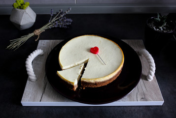 Classical homemade New York cheesecake is lying on dark-violet plate on wooden tray with rope handles on kitchen grey table. Lavender and succulent on background. Red heart to Valentine's day present