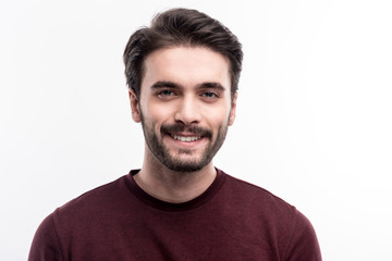 Epitome of masculinity. The portrait of a handsome dark-haired bristled young man in a burgundy sweater smiling at the camera while posing against a white background