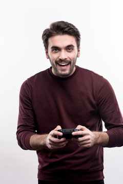 Game lover. Pleasant cheerful young man in a burgundy sweater holding a video game controller while posing against a white background