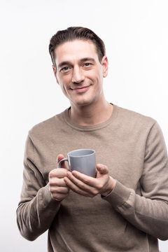 Love this drink. Pleasant handsome man holding a small cup of coffee in his both hands and smiling pleasantly, while posing isolated on a white background