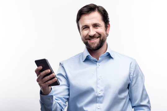 Active user. Cheerful young man posing for the camera and smiling while holding a cell phone in his hand, standing isolated on a white background
