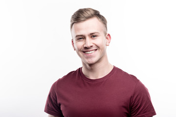 Obraz premium Beautiful man. The portrait of a charming fair-haired young man in a burgundy t-shirt smiling at the camera while posing isolated on a white background