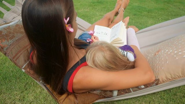 Mother sits with daughter in hammock readng book outdoors