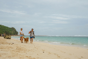 A family of freelancers with a dog is walking along the beach. A stylish young girl leads a gold retriever next to her husband with a surfer.