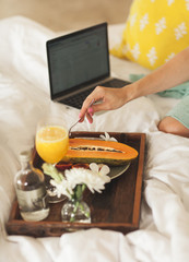 Easy Fruit Breakfast on Tray on White Bad. a Woman's Hand Reaches for Papaya. Laptop on Background