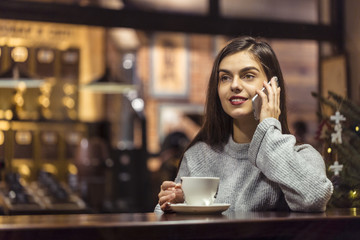 Young pretty brunette girl wears grey sweater have pleasant conversation by smartphone sitting near cafe window