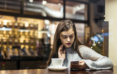 Young pretty brunette girl wears grey sweater browsing internet via smartphone sitting near cafe window
