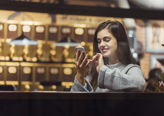 Young pretty brunette girl wears grey sweater browsing internet via smartphone sitting near cafe window