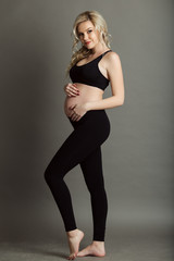 Pregnant beautiful blonde woman is wearing sportive black clothes isolated on gray, healthy life concept