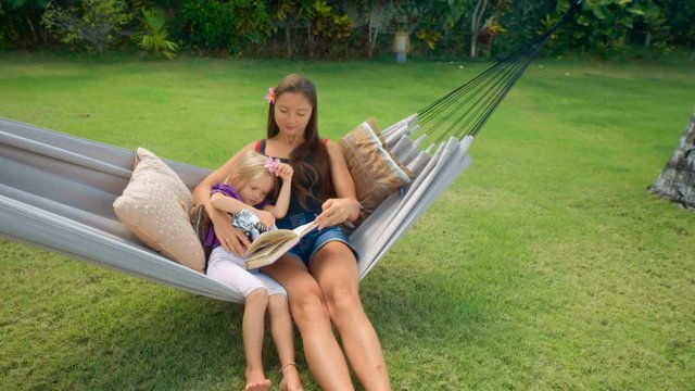 Mother sits with daughter in hammock readng book outdoors