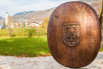 Metal shield with the Coat of arms of Mondsee in Seepromenade Lake promenade Park, St Michael Basilica on the background. Mondsee, Austria