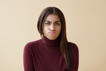 Fototapeta premium Portrait of upset young mixed race woman in maroon turtleneck pouting, feeling disappointed and unhappy after quarrel with her boyfriend, posing in studio with copyspace wall for your advertisement