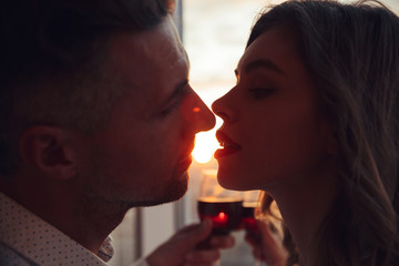 Portrait of lovers kissing at sunset and holding glass with wine at home