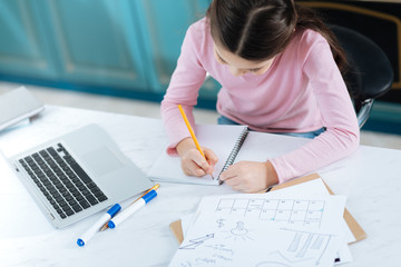 Studying hard. Determined dark-haired little girl writing in her notebook while sitting at the computer and having papers on her table