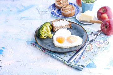 Fried eggs in the form of heart. On a dark plate with broccoli and toast. Blue cup with tea, apples, butter for breakfast and cupcakes. On a light colored background. Free space for text or advertisin