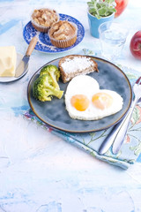 Fried eggs in the form of heart. On a dark plate with broccoli and toast. Blue cup with tea, apples, butter for breakfast and cupcakes. On a light colored background. Free space for text or advertisin