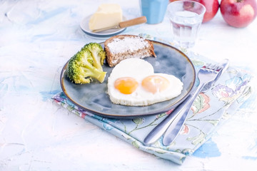 Fried eggs in the form of heart. On a dark plate with broccoli and toast. Blue cup with tea, apples, butter for breakfast. On a light colored background. Free space for text or advertising.