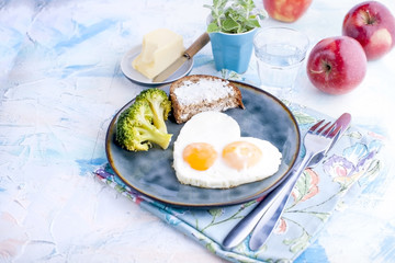 Fried eggs in the form of heart. On a dark plate with broccoli and toast. Blue cup with tea, apples, butter for breakfast. On a light colored background. Free space for text or advertising.