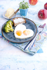 Fried eggs in the form of heart. On a dark plate with broccoli and toast. Blue cup with tea, apples, butter for breakfast. On a light colored background. Free space for text or advertising.